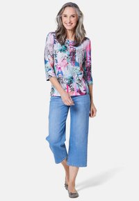 Mehrfarbig bedrucktes Oberteil mit 3/4-Ärmeln, kombiniert mit hellblauen, verkürzten Jeans. Das Oberteil weist florale und abstrakte Muster auf und besteht aus weichem Material.