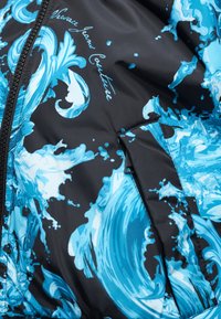 Μαύρο μπουφάν με μπλε και λευκά σχέδια πιτσιλιάς και κυματισμών, φερμουάρ, κάλυμμα τσέπης και επιγραφή "Versace Jeans Couture".