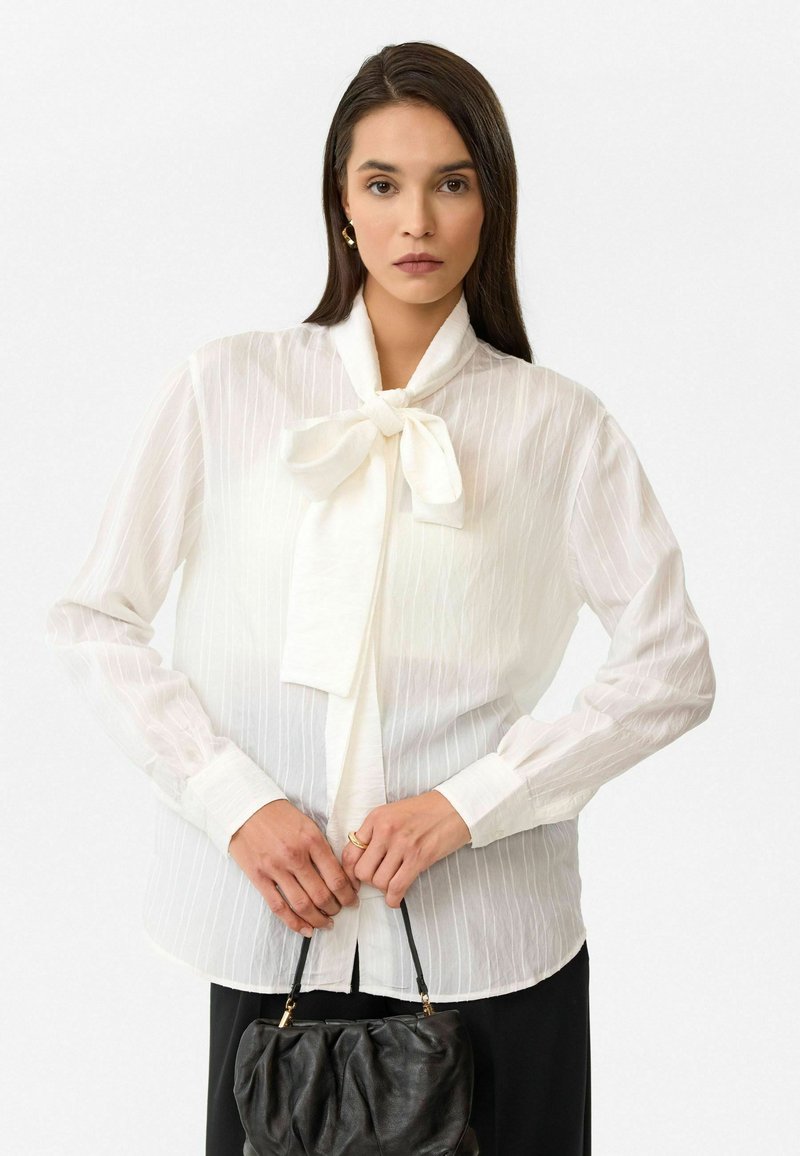 Witte blouse met een strik aan de hals, met verticale strepen, lange mouwen en knoopmanchetten. Gestyled met een zwarte tas.