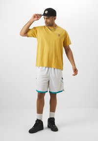 Yellow cotton t-shirt with a logo patch, white mesh shorts with turquoise accents, and black high-top sneakers. Black cap and beaded necklace.