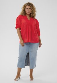 Blouse rouge à manches courtes avec un col en V et des détails froncés, associée à une jupe en jean bleu clair mi-longue avec une fente à l'avant.