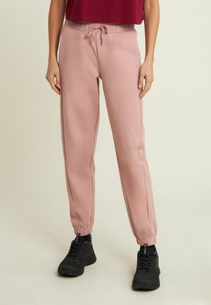 Personne portant un pantalon de jogging rose avec taille à cordon et des baskets noires, se tenant sur un fond clair.