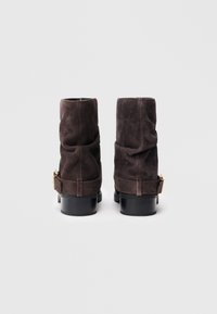 Botas de tobillo de ante marrón con un diseño arrugado, que presentan una correa trasera y herrajes en tono dorado, además de un tacón grueso negro.