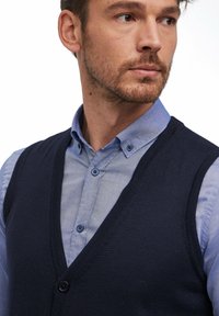 Gilet lavorato a maglia blu navy su una camicia azzurra con bottoni. Il gilet presenta un collo a V e un bottone nero, con una superficie liscia e un design aderente.