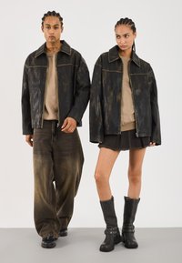Des vestes en cuir noir avec des accents marron clair, coupe oversized, portées sur des tops beiges. Un modèle porte un jean foncé, l'autre une jupe courte et des bottes noires.