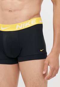 Črne boksarske hlače z rumeno elastiko, na kateri je logo Nike; gladek material s prilegajočim dizajnom in minimalnimi šivi.