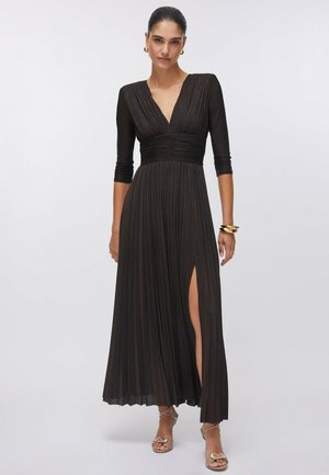 Donna con un lungo vestito nero plissettato con maniche tre quarti e uno spacco alto sulla coscia, che indossa orecchini a cerchio dorati, braccialetti e tacchi con cinturini.