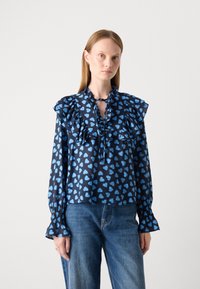 Blusa navy con motivo di cuori blu chiaro, maniche a volant e dettaglio al collo con fiocco. Il tessuto sembra liscio con una leggera lucentezza. Abbinata a jeans blu.