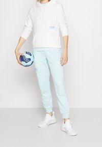 Sudadera blanca con bolsillo tipo canguro y logo azul, combinada con pantalones de chándal azul claro. El atuendo se completa con zapatillas deportivas blancas y una pelota multicolor.