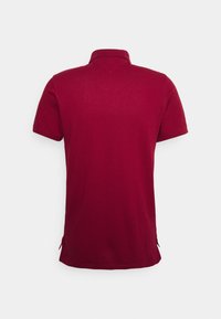 Burgundin lyhythihainen polo-paita, joka näkyy takaa, kauluksella ja sivuhalkioilla helman kohdalla.