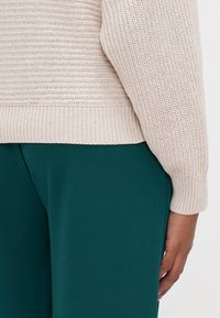 Μπεζ πλεκτό πουλόβερ με ριμπ μανίκια και στρίφωμα, συνοδεύεται από πράσινα παντελόνια από λείο ύφασμα, που αναδεικνύουν επιφάνεια με υφή και εφαρμοστή σχεδίαση.