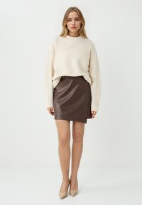 Maglione beige a maglia con scollo tondo, abbinato a una gonna aderente in finta pelle marrone. La modella indossa décolleté beige con punta appuntita.