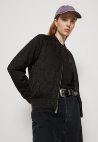 Veste bomber noire avec texture florale, poignets côtelés, fermeture éclair à l'avant, associée à un t-shirt noir, un jean foncé et une casquette lavande.