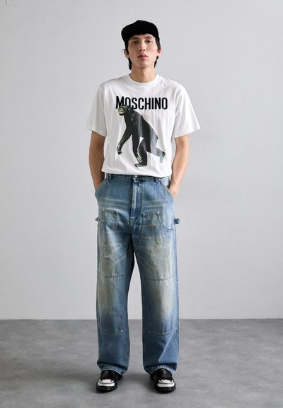 Hvid t-shirt med sort grafisk print og teksten "MOSCHINO", parret med lyseblå slidte brede jeans og sorte sko.