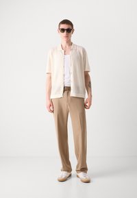 Chemise beige clair à manches courtes avec des rayures verticales, associée à un pantalon beige et des baskets blanches. Comprend des lunettes de soleil rondes et une ceinture fine.