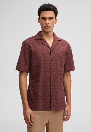 Junger Mann mit lockigem Haar, der ein strukturiertes, kurzärmeliges, bordeauxrotes Hemd mit Knöpfen und beigefarbene Hose vor einem einfarbigen Hintergrund trägt.