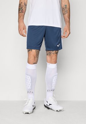 Navy athletic shorts, white jersey, white knee-high socks with grey text, and white cleats with black accents. Tattoos on visible skin.