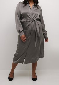 Robe chemise grise en tissu soyeux avec manches longues, fermeture à boutons sur le devant et détail de taille nouée. Caractérisée par un ourlet à hauteur des genoux et une texture lisse.