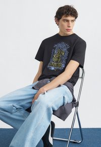 Νέος άνδρας με σγουρά μαλλιά κάθεται σε μεταλλική καρέκλα, φορώντας μαύρο γραφικό μπλουζάκι Puma, ανοιχτόχρωμα τζιν και μαύρα-λευκά αθλητικά παπούτσια.