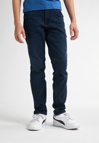 Jeans de mezclilla azul oscuro con corte ajustado, que cuentan con cinco bolsillos y un desvanecimiento sutil. Llevados con zapatillas blancas y una camiseta azul.