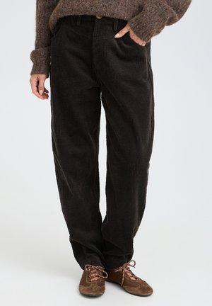 Personne portant un pantalon ample en velours côtelé foncé, un pull marron texturé et des chaussures décontractées marron, debout devant un fond uni.