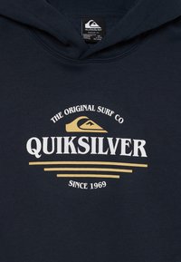 Tengerészkék kapucnis pulóver, pamutból készült, fehér és arany Quiksilver logóval. Jellemzője egy kenguruzseb és bordázott mandzsetták.
