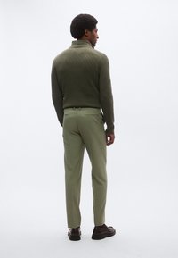 Mørkegrøn ribstrikket rullekravesweater, lysegrøn skræddersyet bukser, sorte sko, set bagfra. Glatte teksturer og tætsiddende design.