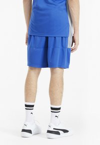 Pantaloni sportivi blu a due strati, con cintura elastica e pannelli laterali a contrasto. Sneakers bianche con dettagli neri e calze a strisce.