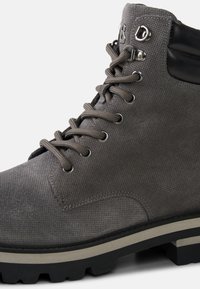 Šedá turistická bota s texturovaným svrškem, černým polstrovaným límcem, kovovými očky a silnou gumovou podrážkou s drážkami pro lepší trakci.