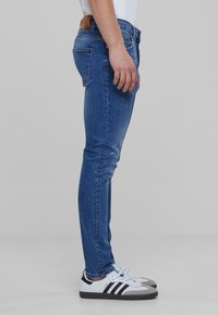 Jeans skinny azul con ligero desgaste, material de algodón, diseño de cinco bolsillos y etiqueta de cuero marrón en la cintura, combinados con zapatillas blancas y negras.