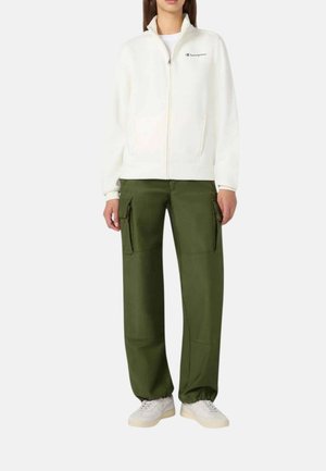 Personne portant une veste polaire blanche zippée avec le logo Champion, un pantalon cargo vert avec des poches, et des baskets blanches.