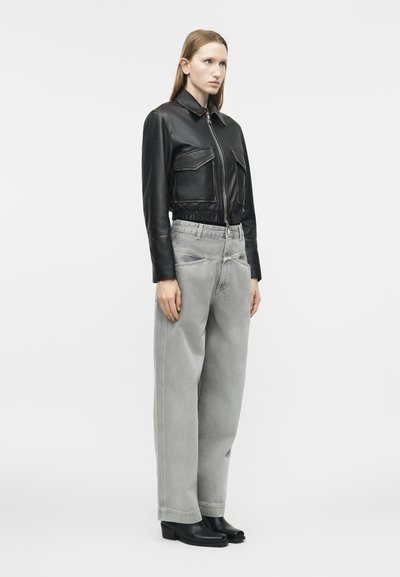 Γυναίκα που στέκεται φορώντας μια μαύρη κοντή δερμάτινη ζακέτα, φαρδιά ανοιχτό γκρι τζιν και μαύρες μποτάκια αστραγάλου σε ένα απλό λευκό φόντο.
