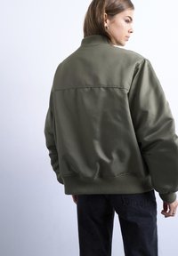 Femme portant une surchemise bomber vert olive oversize et un jean noir, debout de profil sur un fond blanc uni.