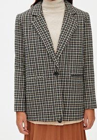 Blazer a quadri con un motivo nero, bianco e beige, chiusura con un bottone singolo e due tasche frontali, indossato su un dolcevita color crema.