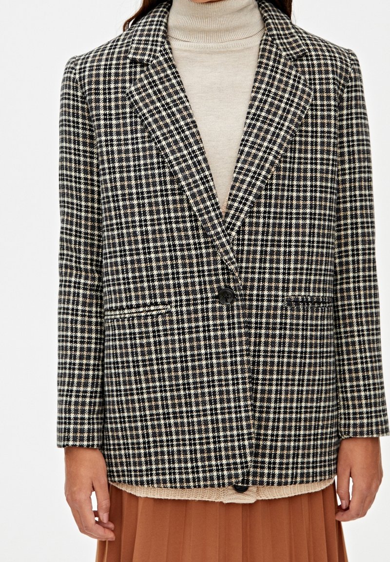 Blazer a quadri con un motivo nero, bianco e beige, chiusura con un bottone singolo e due tasche frontali, indossato su un dolcevita color crema.