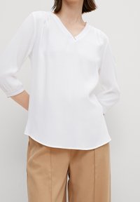 Blusa bianca a maniche lunghe con scollatura a V e dettagli plissettati sulle spalle. Tessuto morbido e leggero con orlo arrotondato, abbinata a pantaloni color sabbia.