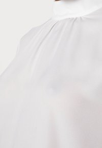 Blouse blanche à col montant en tissu lisse, présentant de délicats plis le long du col et une texture légère et fluide.