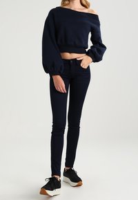 Kvinna som bär en off-shoulder svart cropped tröja, svarta skinny jeans och svarta plattformssneakers med vita sulor, stående mot en enfärgad bakgrund.