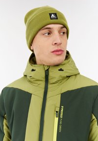 Zelena i tamnozelena jakna s visokim ovratnikom, patentnim zatvaračem sprijeda i detaljima na džepovima. Uz to ide rebrasti zeleni šešir s logom.