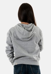 Persona con cabello largo y oscuro, vistiendo una sudadera gris claro y jeans azul oscuro, vista desde atrás sobre un fondo liso.