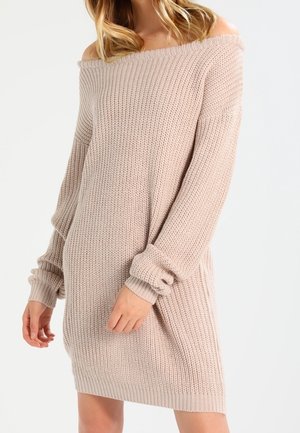 Femme portant une robe pull en tricot beige clair, épaules dénudées, manches longues avec un motif texturé, debout devant un fond uni.