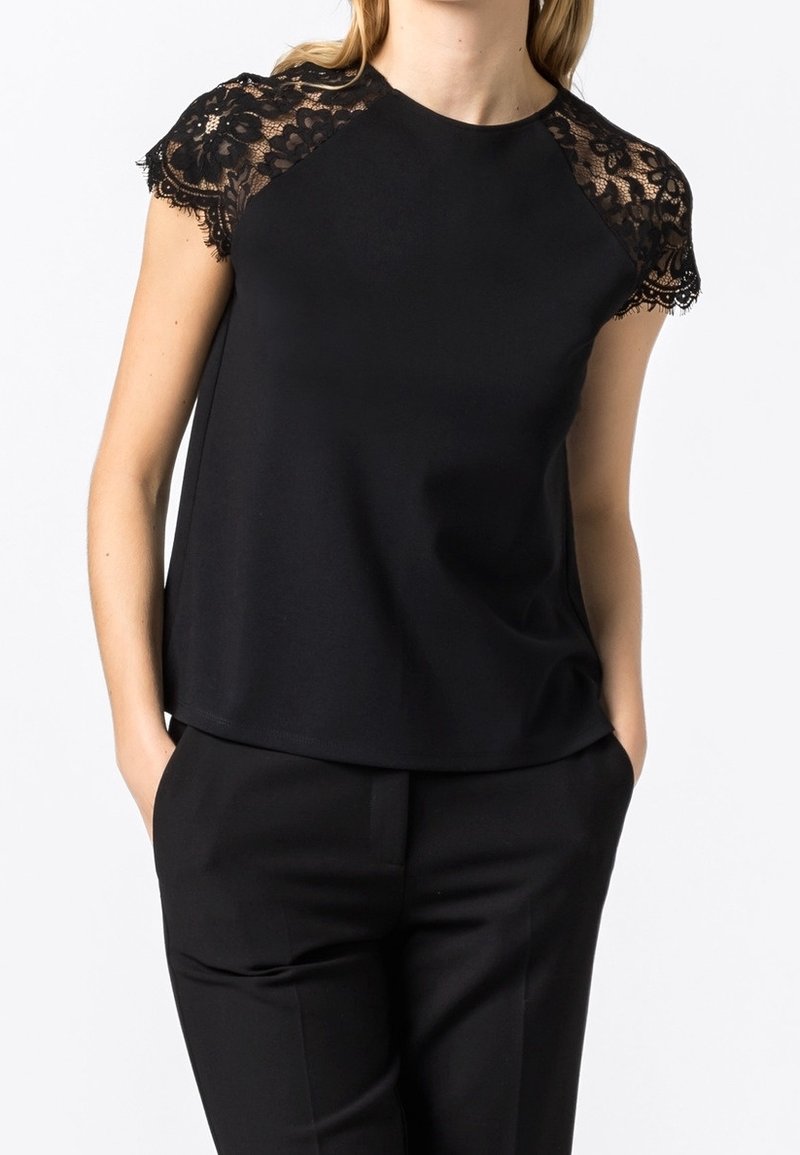 Femme portant un haut noir à manches courtes avec un détail en dentelle aux épaules et un pantalon noir, mains dans les poches, sur fond blanc.