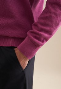 Maglione viola con tessuto a trama e polsini a costine, abbinato a pantaloni scuri. La mano è appoggiata nella tasca dei pantaloni.