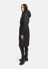 Manteau rembourré noir avec capuche, taille ceinturée, poches latérales zippées et design matelassé. Associé à un jean foncé et des bottes noires à lacets.