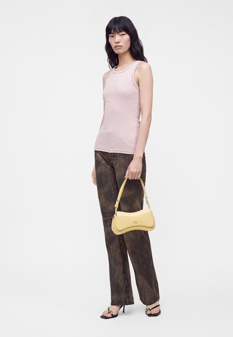 Femme portant un top sans manches rose clair, un jean marron foncé, des sandales noires à talons, tenant un petit sac à main jaune sur un fond blanc uni.