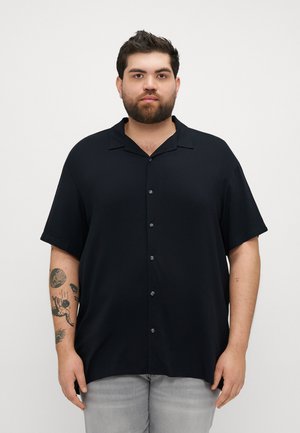 Chemise noire à manches courtes avec un col évasé, texture lisse et coupe décontractée. Pantalons en denim gris visibles sous la chemise.