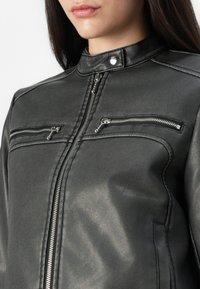 Černá faux kožená bunda s vysokým límcem, dvěma kapsami na zip na hrudi a elegantními detaily šití. Hladká textura a moderní design.