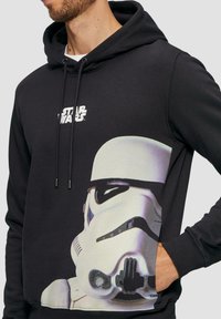 Czarna bluza z kapturem z białym logo "Star Wars" oraz graficznym nadrukiem hełmu Stormtroopera z przodu. Wykonana z gładkiego materiału.