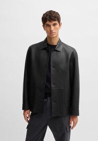 Black leather jacket with a collared neck, front buttons, two side pockets, and a smooth texture. Paired with grey pinstriped trousers.