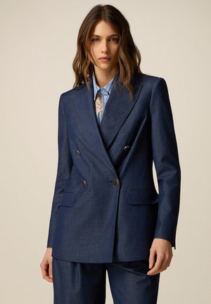 Donna che indossa un blazer doppiopetto blu navy su misura con bottoni argentati sopra una camicia a righe blu con una sciarpa fantasia.